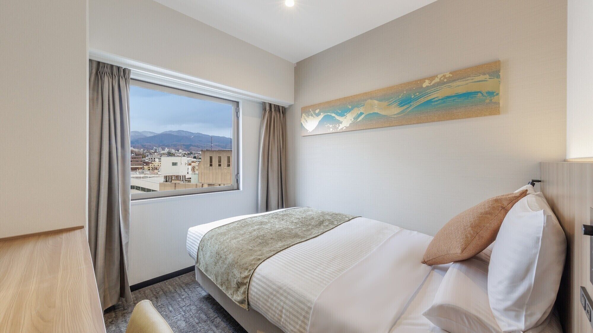 A hotel room with a bed, a window with curtains, a painting on the wall, and a view of buildings and mountains.