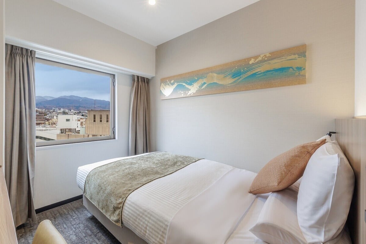 A hotel room with a bed, a window with curtains, a painting on the wall, and a view of buildings and mountains.