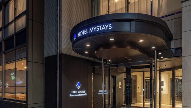 L'entrée de l'hôtel Mystays se distingue par son design moderne, avec une porte d'entrée en verre et une enseigne placée au-dessus de la porte.