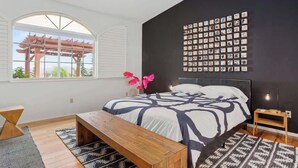 Une chambre avec un lit, un banc, un mur orné de photos encadrées et une fenêtre donnant sur une pergola.