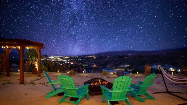 Une scène nocturne avec un ciel étoilé, un foyer et des chaises vertes disposées sur une zone sablonneuse.
