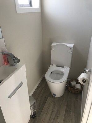 A small, clean bathroom with a white toilet, a trash can, a wicker basket with toilet paper, and a window above the toilet.