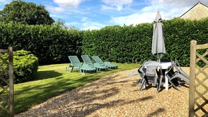 A patio area with outdoor seating, including lounge chairs and a table with chairs, surrounded by a hedge and a gravel path.