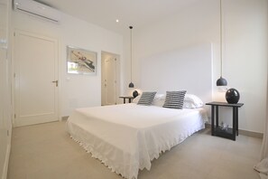 A bedroom with a large bed, a bedside table with a lamp, a door, and a wall-mounted artwork.