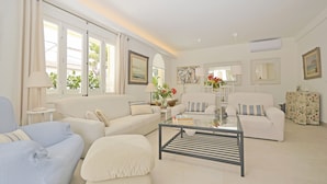 Un salon spacieux avec un canapé, un pouf et une table basse, de grandes fenêtres avec des rideaux et un tableau accroché au mur.