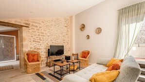 Un salón con pared de piedra, un sofá gris con cojines amarillos, un televisor sobre un mueble bajo, dos sillas de mimbre y una ventana con cortinas.