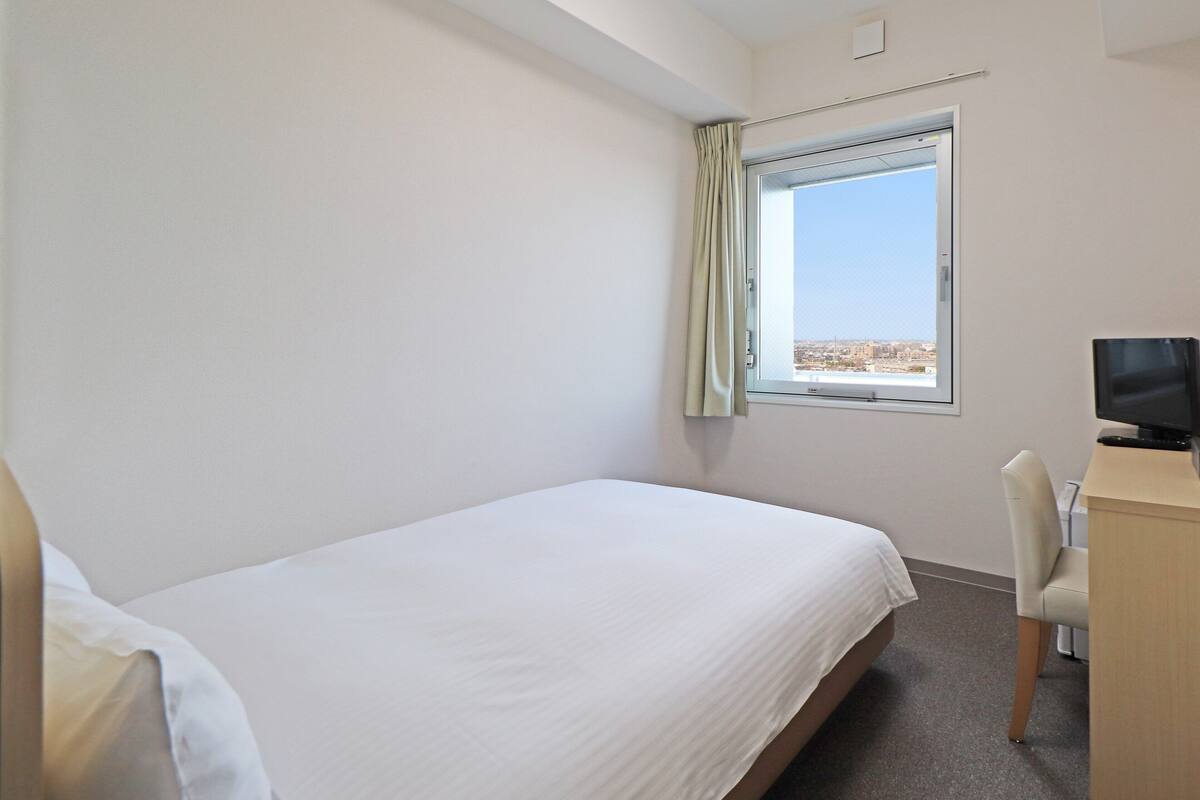 Une chambre d’hôtel avec un lit simple, un bureau avec un ordinateur et une fenêtre donnant sur la ville.