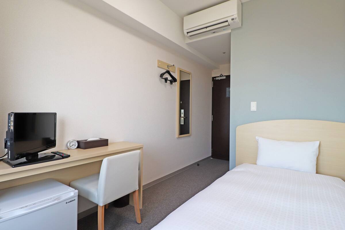 Une chambre d’hôtel comprenant un lit, un bureau avec une télévision, une chaise, un petit réfrigérateur et un appareil de climatisation.