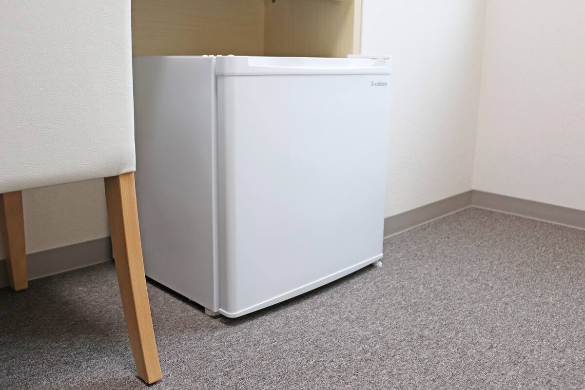 Un petit réfrigérateur blanc, placé dans un coin d’une pièce dont le sol est en bois et un des murs est blanc.