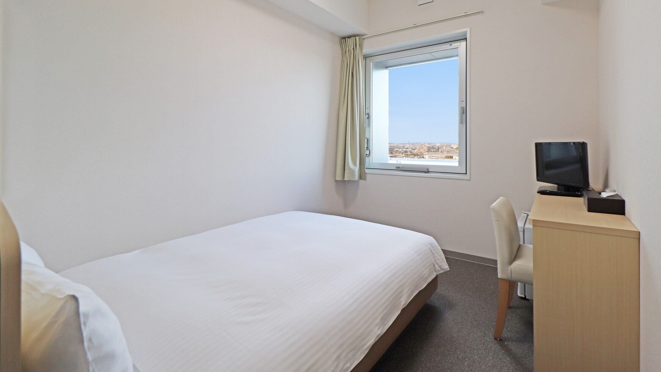 Ein Hotelzimmer mit einem Einzelbett, einem Schreibtisch mit Computer, einem Sessel und einem Fenster mit Blick auf die Stadt.