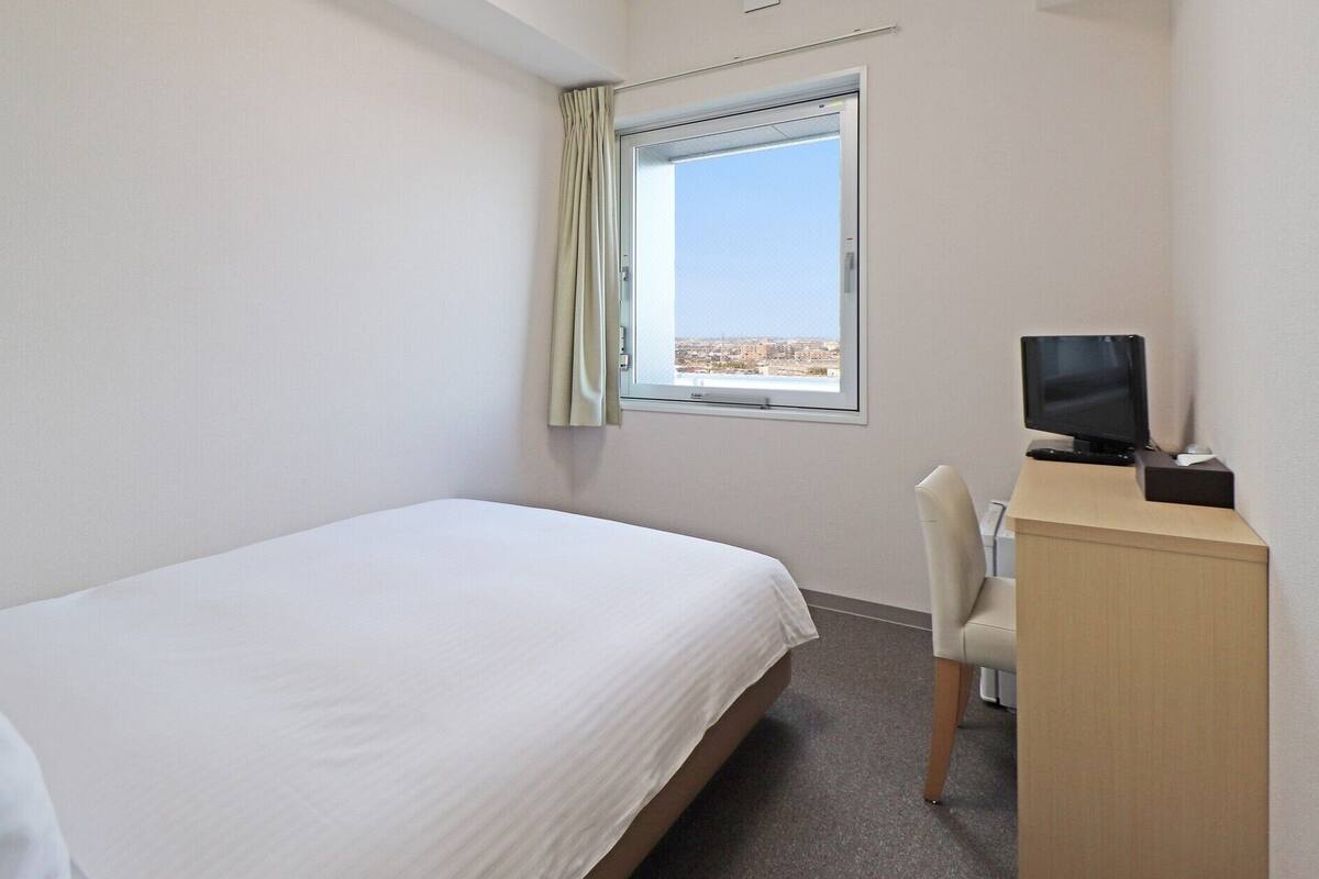 Une chambre d’hôtel avec un lit simple, un bureau avec un ordinateur, une chaise et une fenêtre donnant sur la ville.