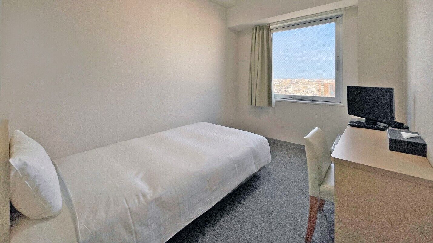 Ein Hotelzimmer mit einem Einzelbett, einem Schreibtisch mit Computer und einem Fenster mit Blick auf die Stadt.