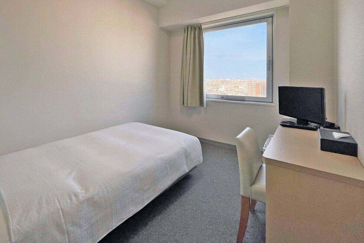 Une chambre d’hôtel avec un lit simple, un bureau avec un ordinateur et une fenêtre donnant sur la ville.