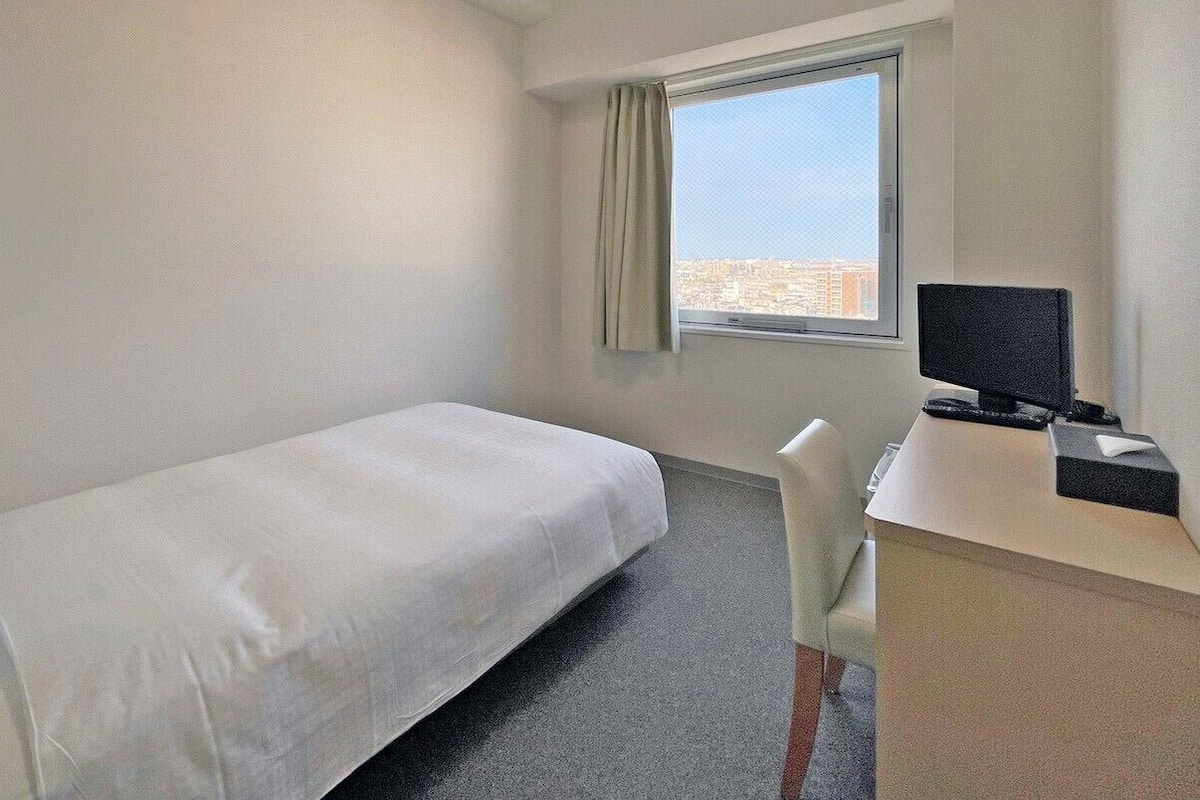A hotel room with a single bed, a desk with a computer, and a window with a city view.