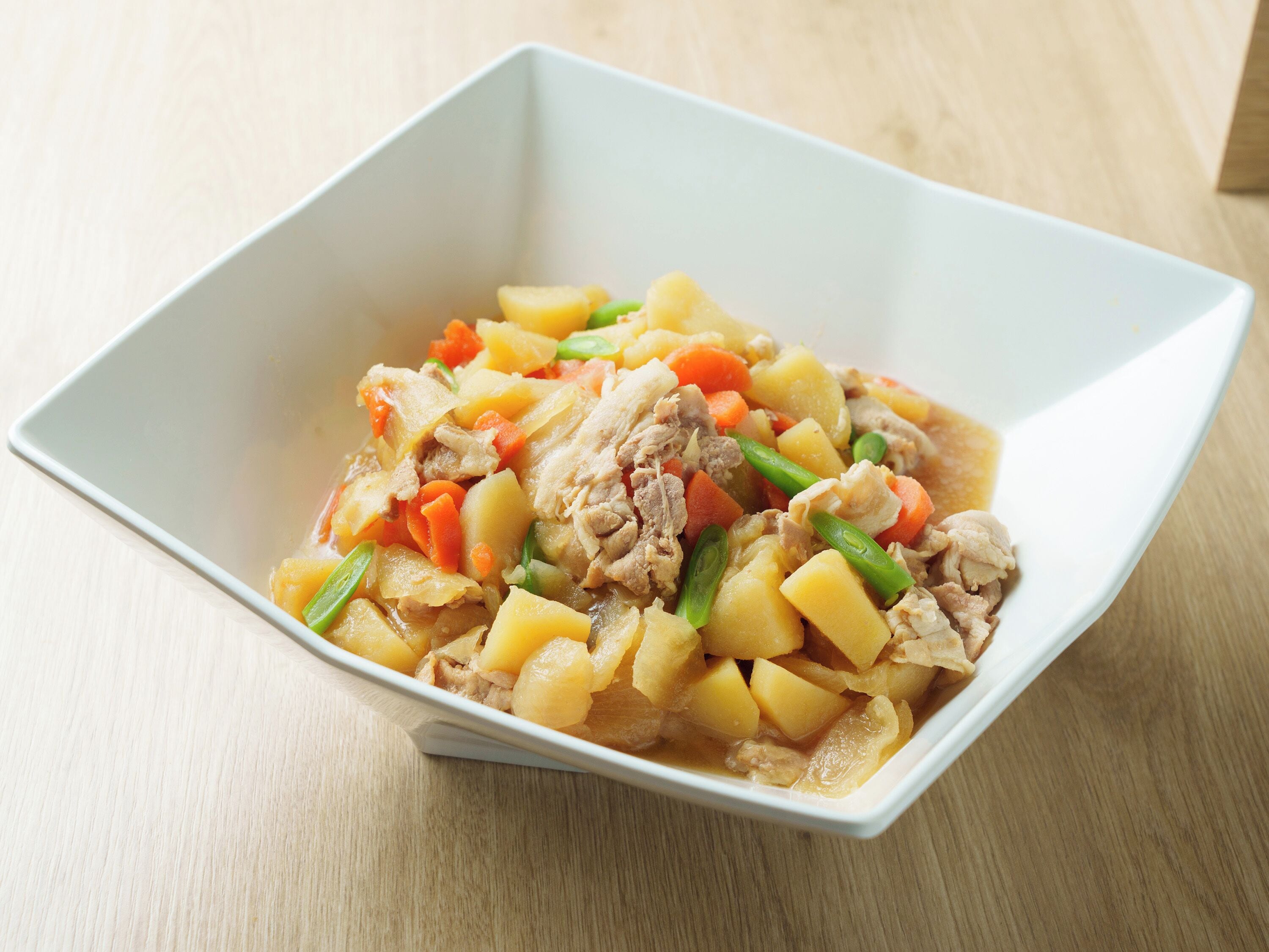A white square bowl filled with a stew containing chunks of meat, potatoes, carrots, and green onions.