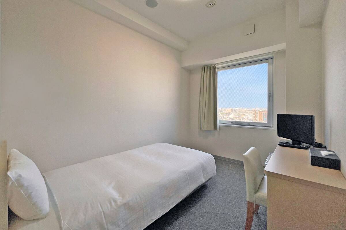 Une petite chambre propre, avec un lit simple, un bureau équipé d’un ordinateur et une fenêtre donnant sur la ville.