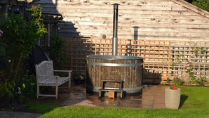 A wooden structure with a hot tub, a bench, and a small table in a garden setting. -  5 Star luxury barn in the Heart of England forest with wood-fired Hot tub. (Great Alne)