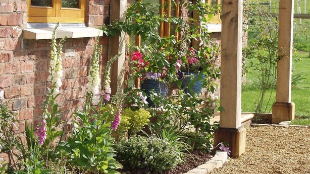 A garden with various plants and flowers in front of a brick building with a window and wooden beams. -  5 Star luxury barn in the Heart of England forest with wood-fired Hot tub. (Great Alne)