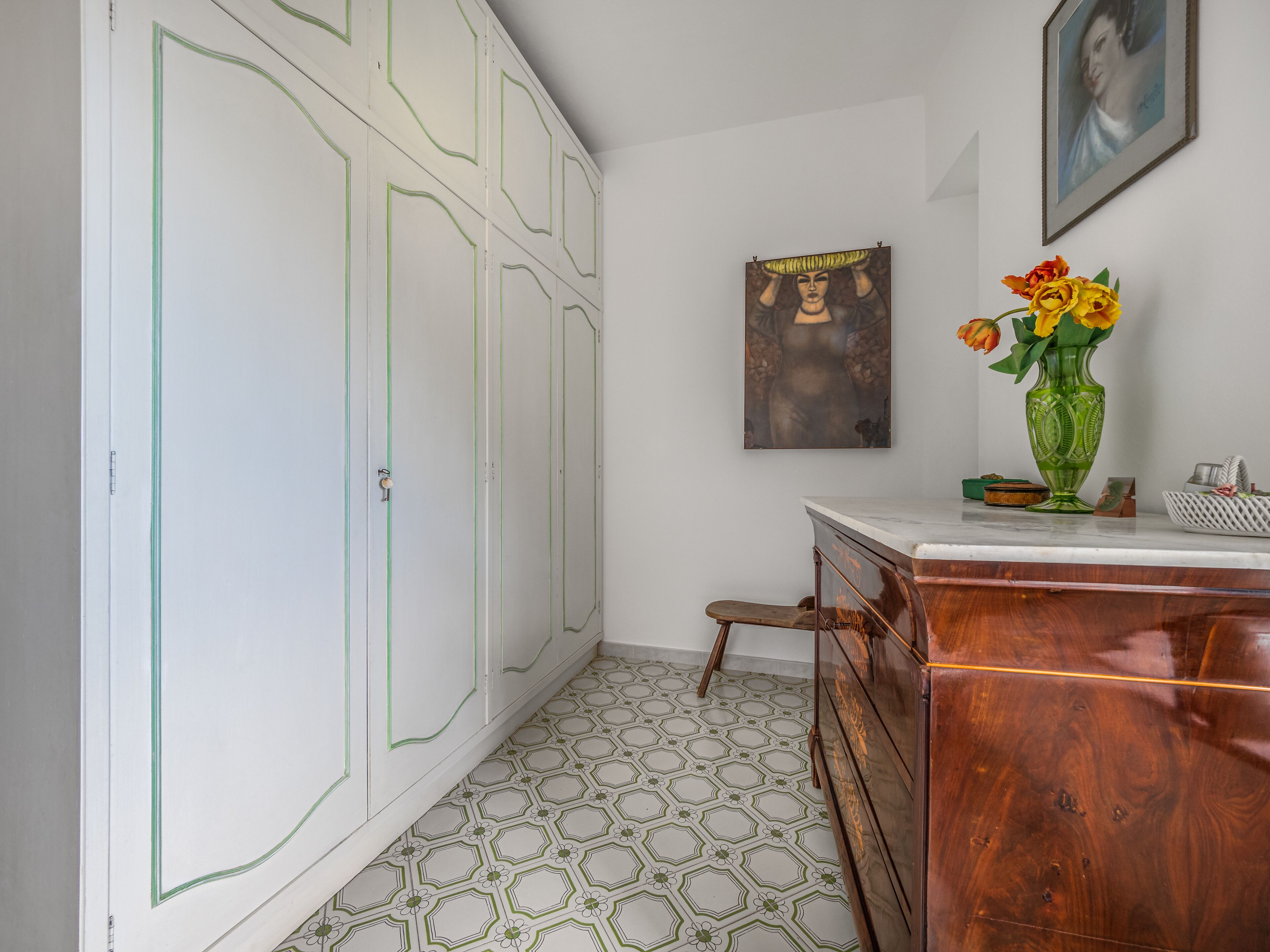 A room with a wooden dresser, a green vase with flowers, a framed picture on the wall, and patterned floor tiles.