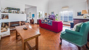 A spacious living room with a red sofa, a green armchair, and a wooden table. There is a fireplace, a balcony with a view, and various decorative items on the walls. - Casa Lauro (Positano)