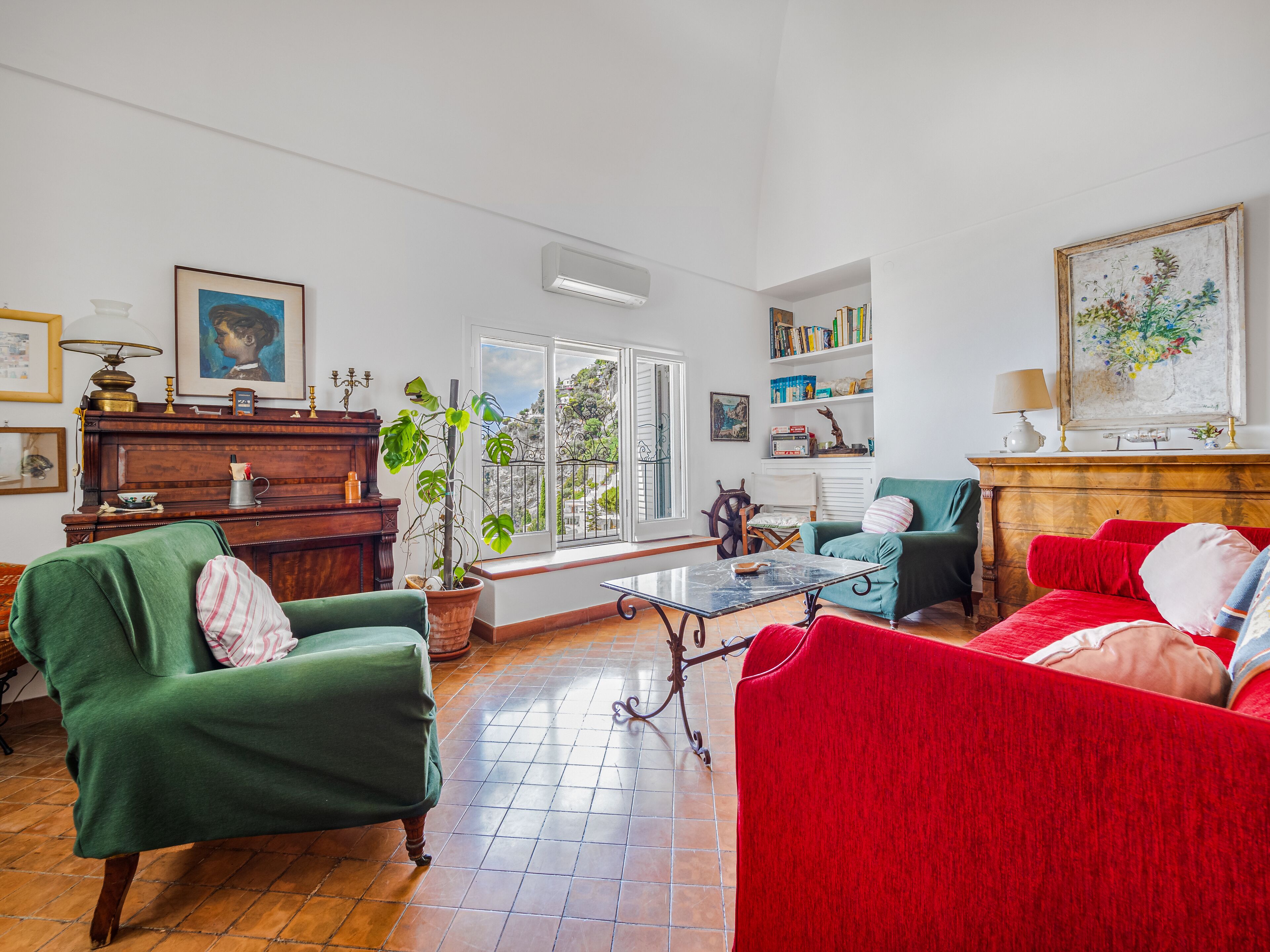 Un salón amplio con un piano, sofás verde y rojo, una mesa de centro y un ventanal con vistas a una colina.