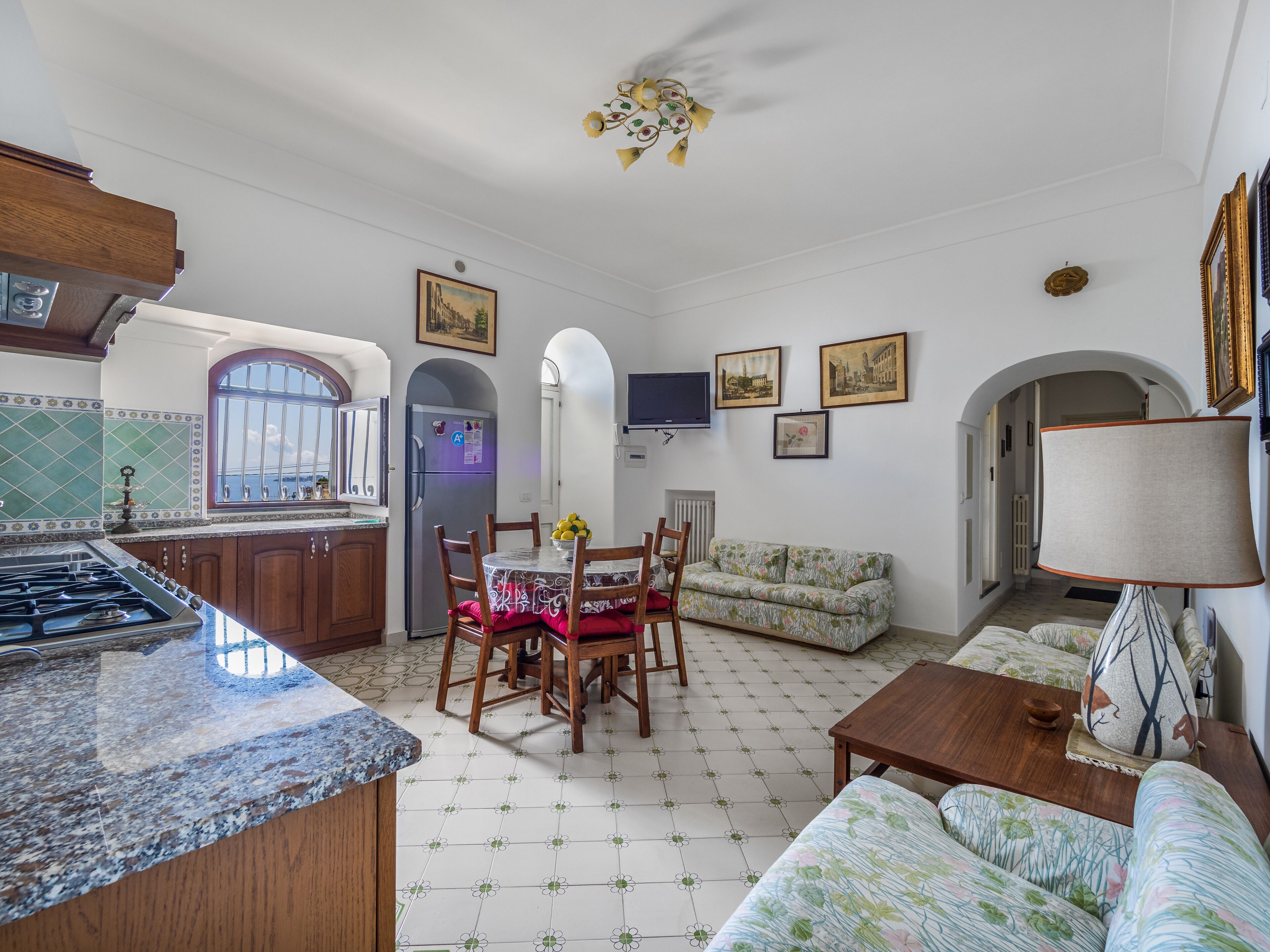 A spacious living area with a kitchen, dining table, and sofa. There is a TV mounted on the wall and a ceiling fan.