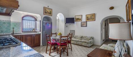 A spacious living area with a kitchen, dining table, and sofa. There is a TV mounted on the wall and a ceiling fan.