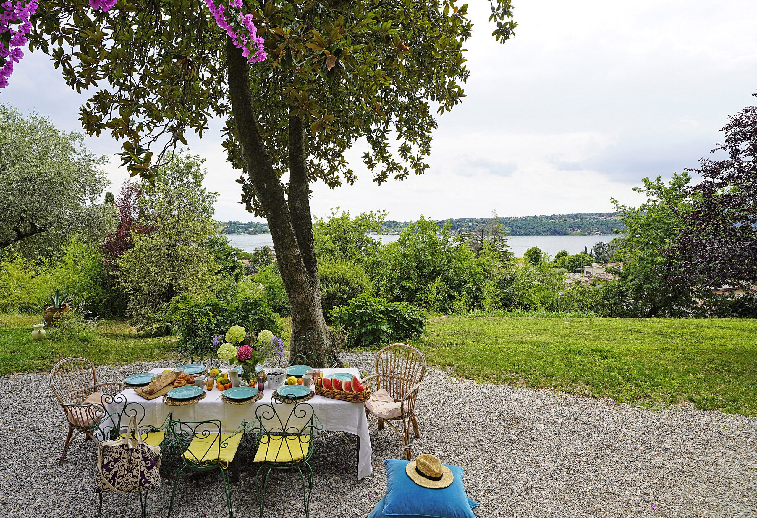 An outdoor dining setup with a table set for four, surrounded by chairs, under a tree with a view of a lake.