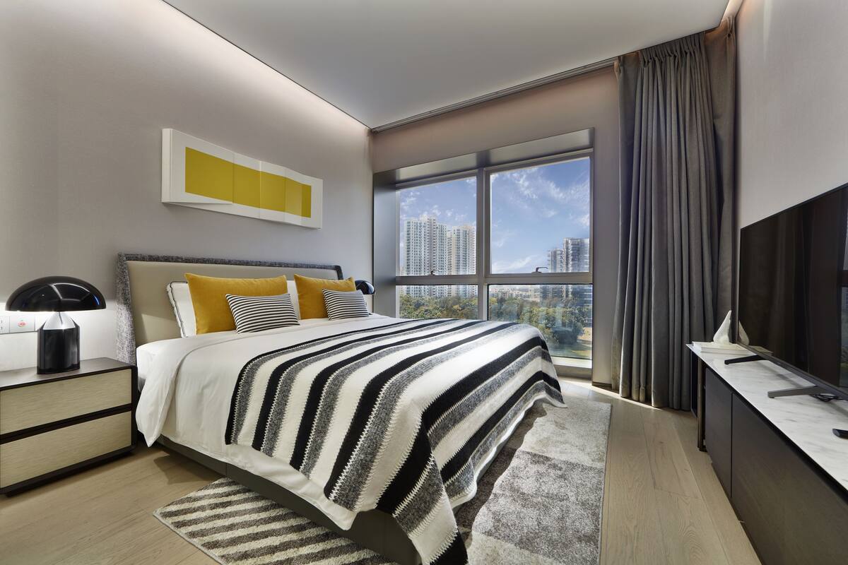 A modern bedroom with a large bed, a nightstand, a desk, and a view of high-rise buildings.