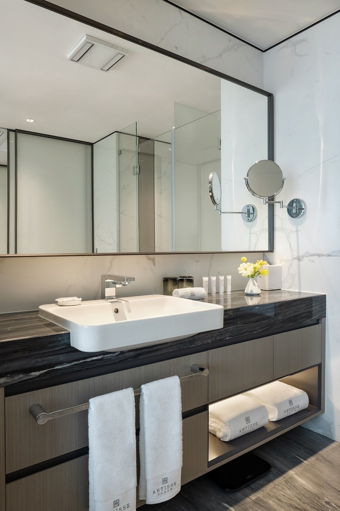 A modern bathroom with a large mirror, white sink, and marble countertop. There are towels and toiletries on the counter.