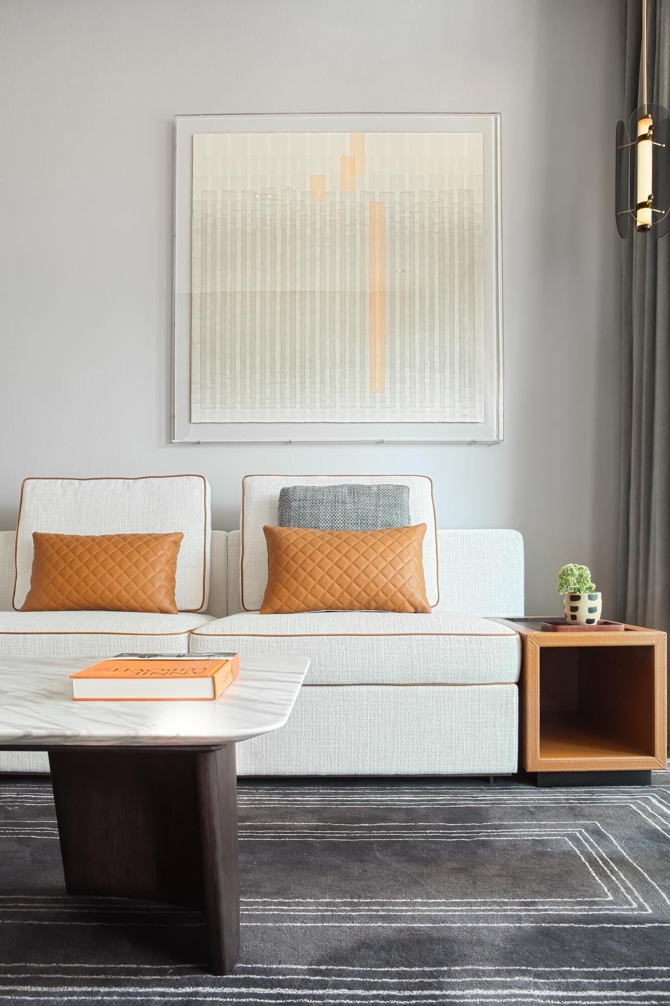 A modern living room with a sofa, two pillows, a coffee table, a framed abstract artwork, a side table with a plant, and a dark rug.