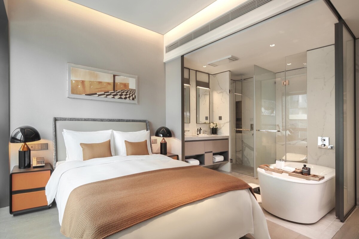 A modern hotel room with a large bed, a bathroom with a glass shower enclosure, and a small sitting area.