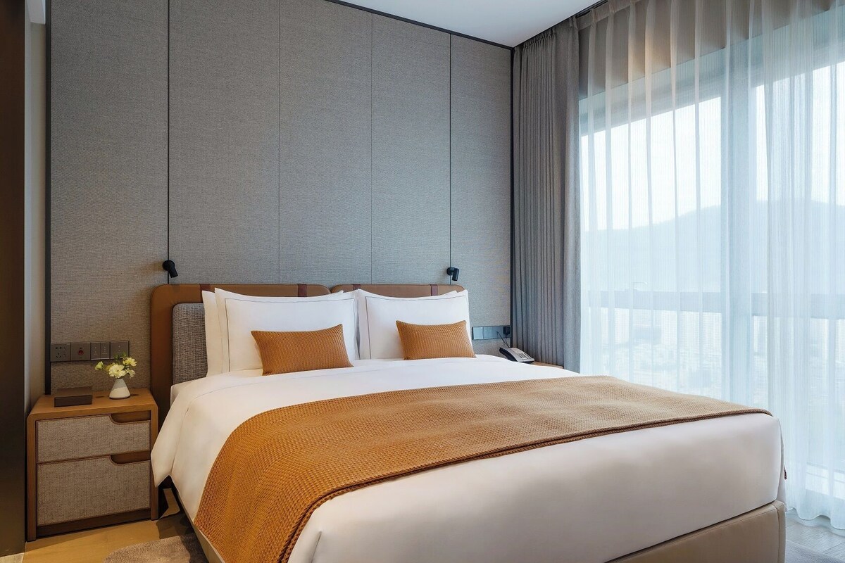 A hotel room with a large bed, a bedside table with a lamp, and a view of the city through sheer curtains.