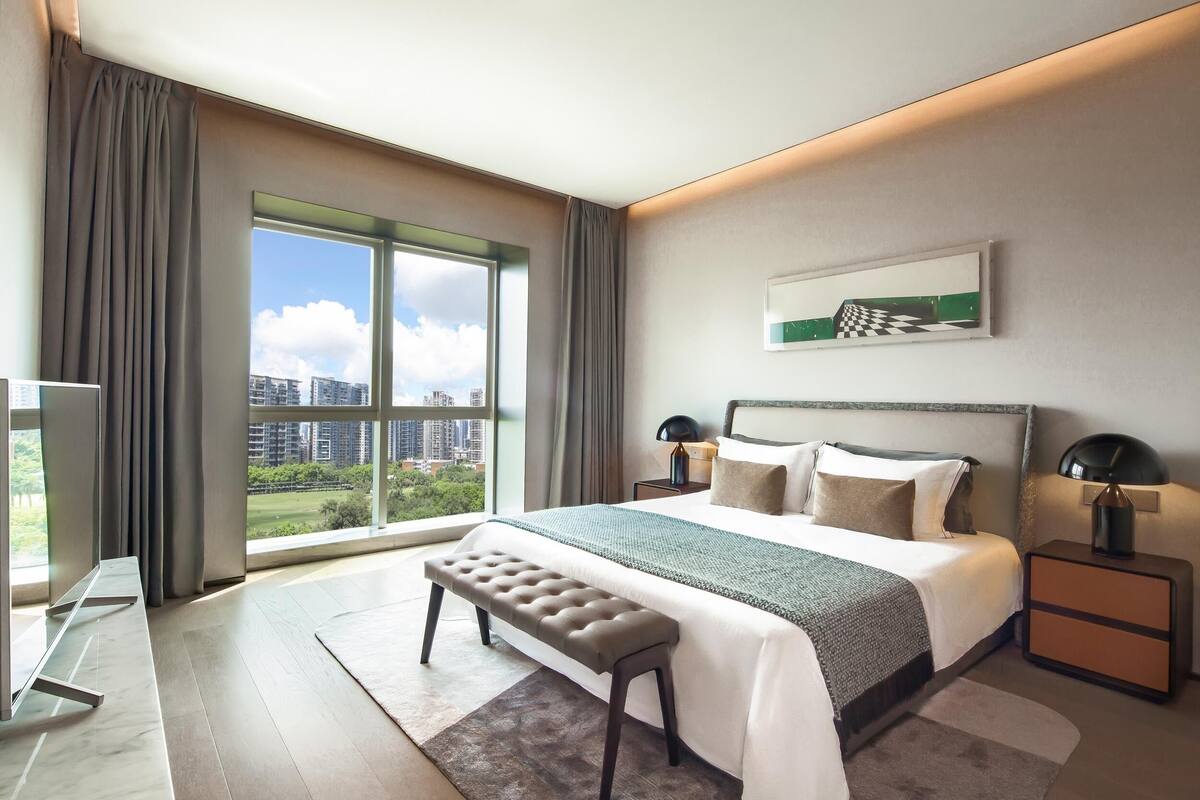 A modern hotel room with a large bed, a bench, a desk, and a view of the city through large windows.
