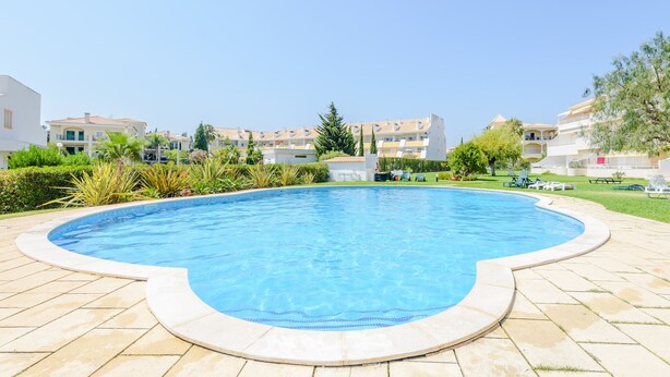 Une piscine dotée d’une terrasse carrelée, entourée de verdure et avec des bâtiments modernes en arrière-plan.