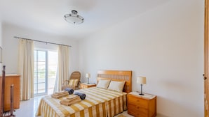 Une chambre à coucher avec une couvre-lit à carreaux, une tête de lit en bois, une table de chevet avec une lampe, une chaise et une vue sur l’extérieur par une fenêtre ornée de rideaux.