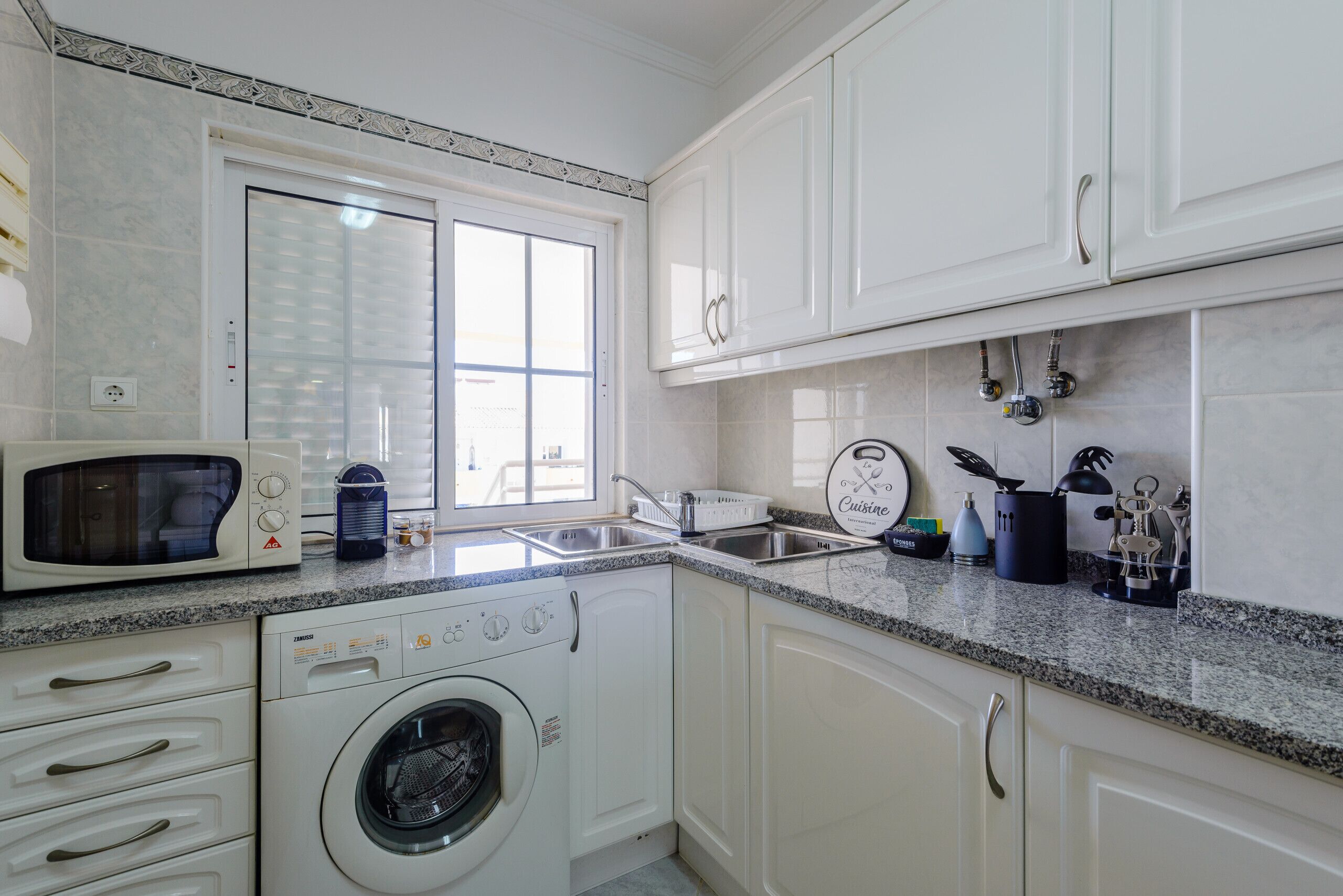 A kitchen with white cabinets, a built-in microwave, a washing machine, and a sink with a window above it.