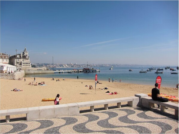 A beach with people sunbathing and swimming, a pier with boats, and a historic building in the background.