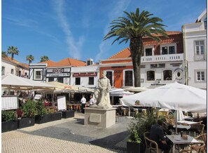 A plaza with a statue, outdoor seating, and various buildings with signs indicating restaurants and cafes.