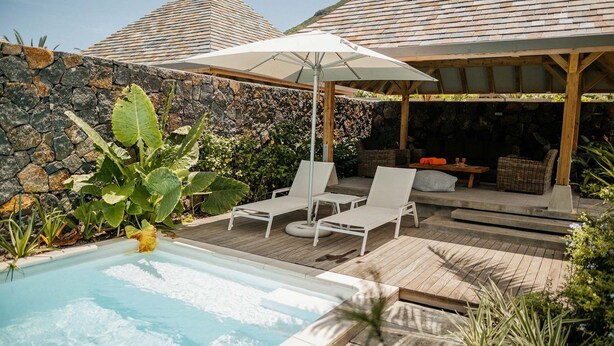 Een zwembad met ligbedden en parasols, omringd door een stenen muur en weelderige begroeiing.
