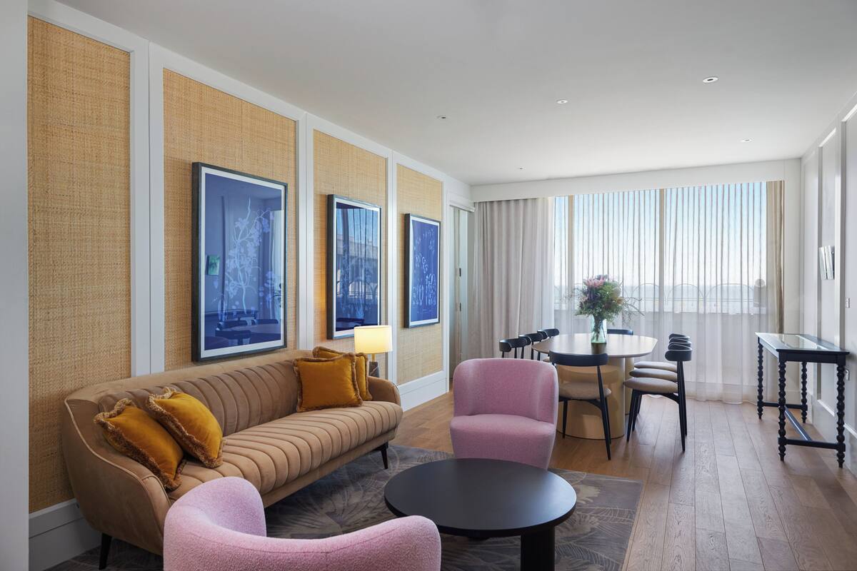 Een moderne hotelkamer met een bank, fauteuils, een eettafel en een groot raam met gordijnen.
