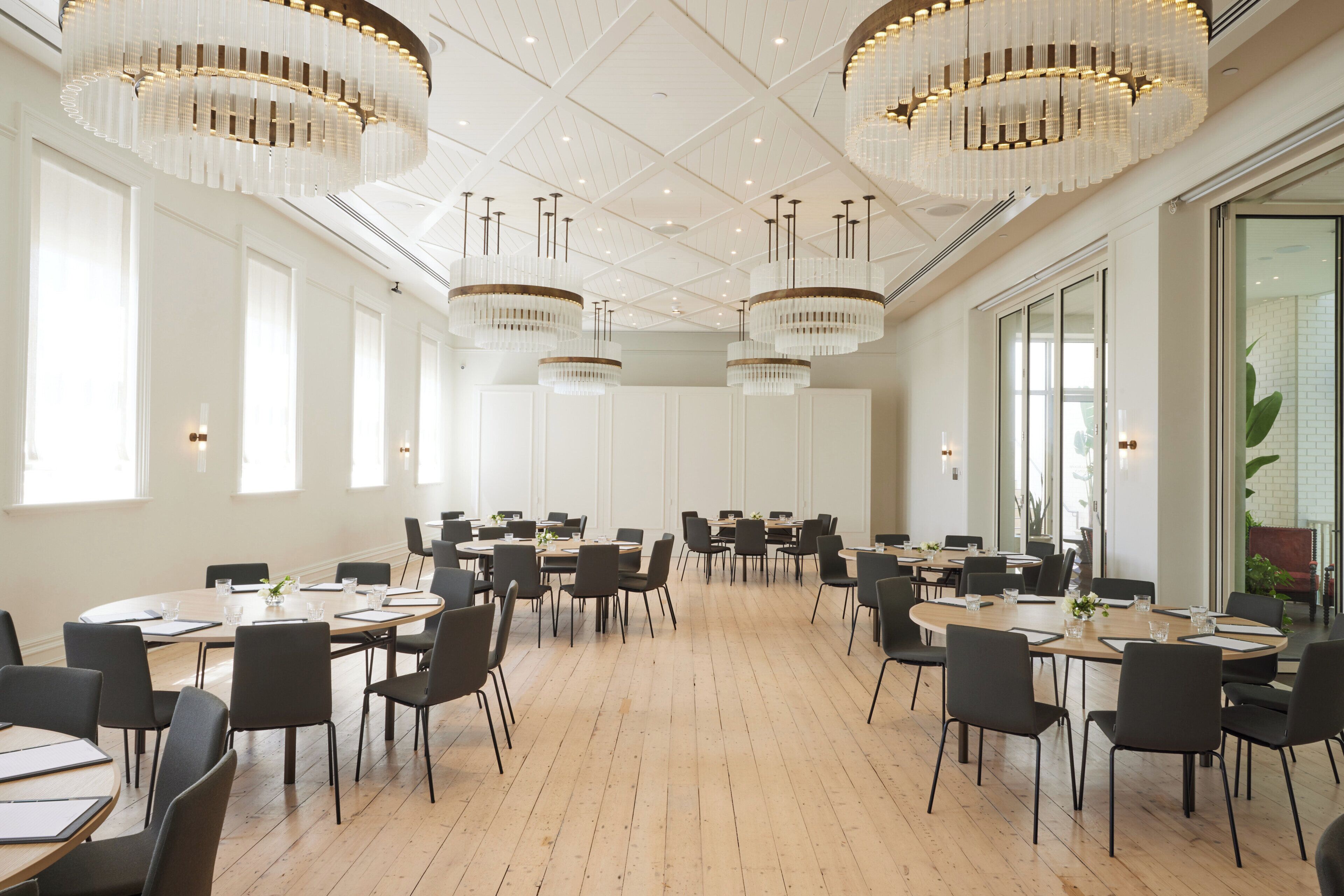 A spacious conference room with multiple tables and chairs, large windows, and elegant chandeliers.