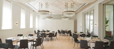 A spacious conference room with multiple tables and chairs, large windows, and elegant chandeliers.