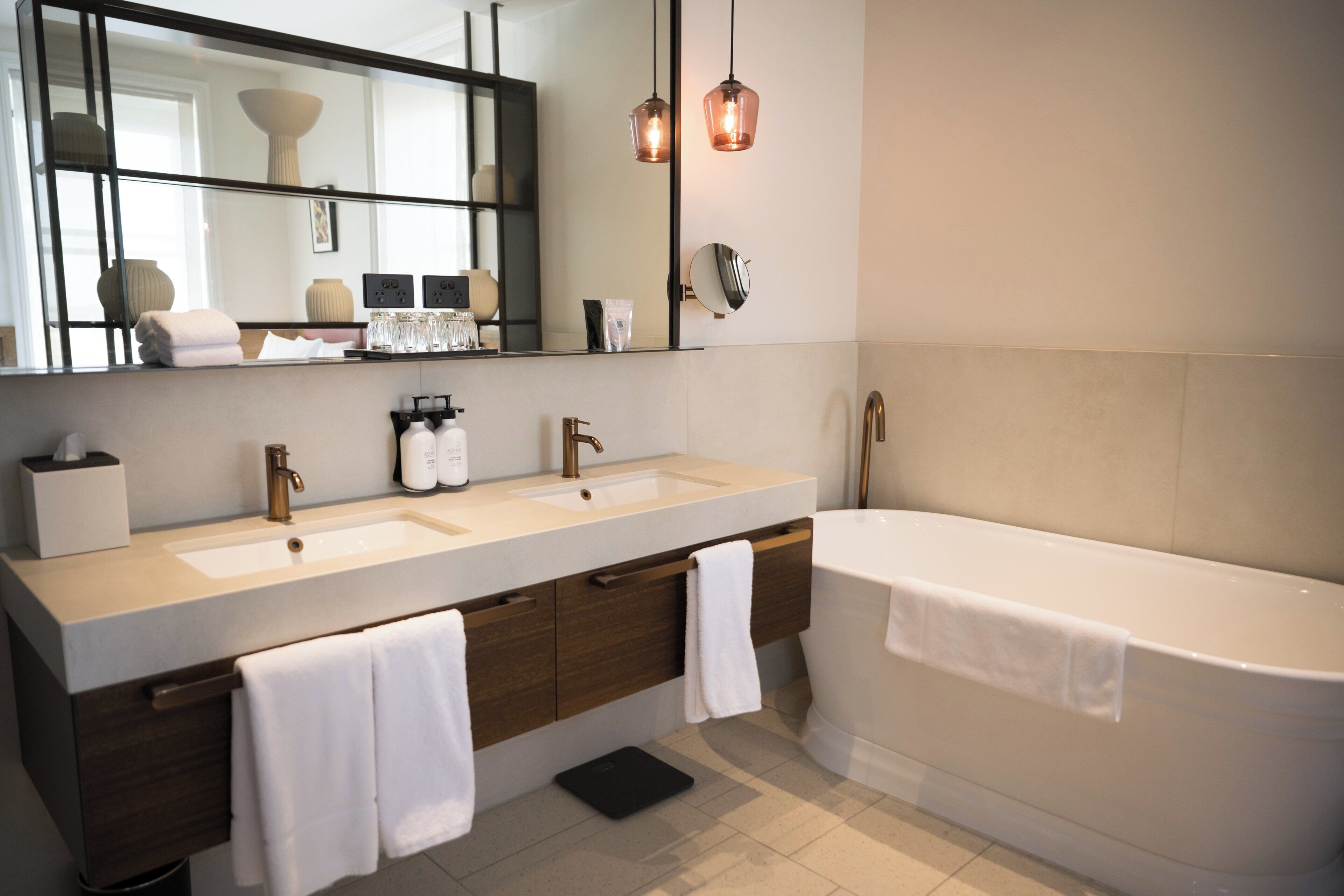 Un baño moderno con dos lavamanos, un espejo grande y una bañera. Hay toallas colgadas al lado de la bañera y una balanza negra en el suelo.