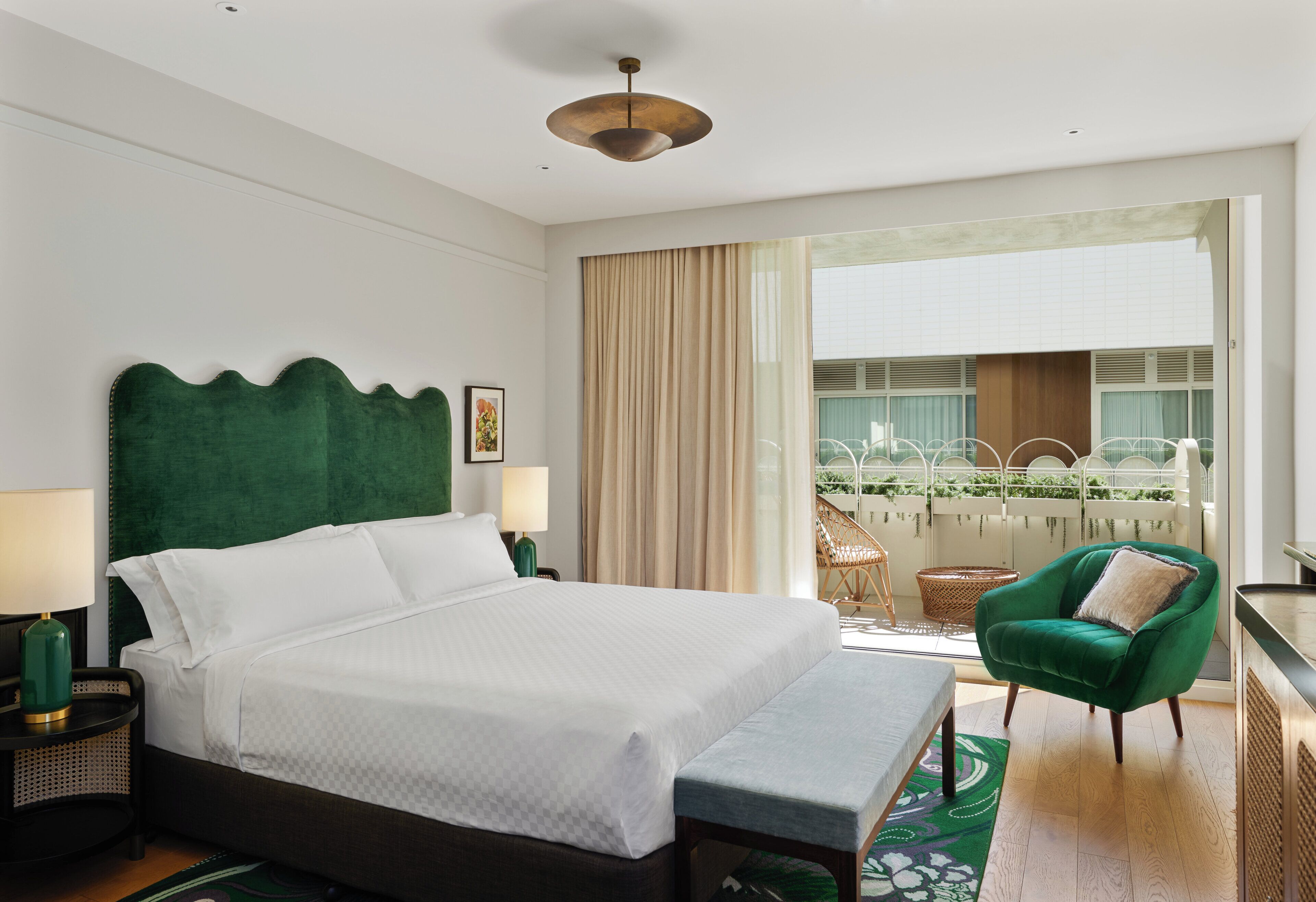Habitación de hotel con una cama grande, un sillón verde, un balcón con vistas a zonas verdes y una lámpara en el techo.