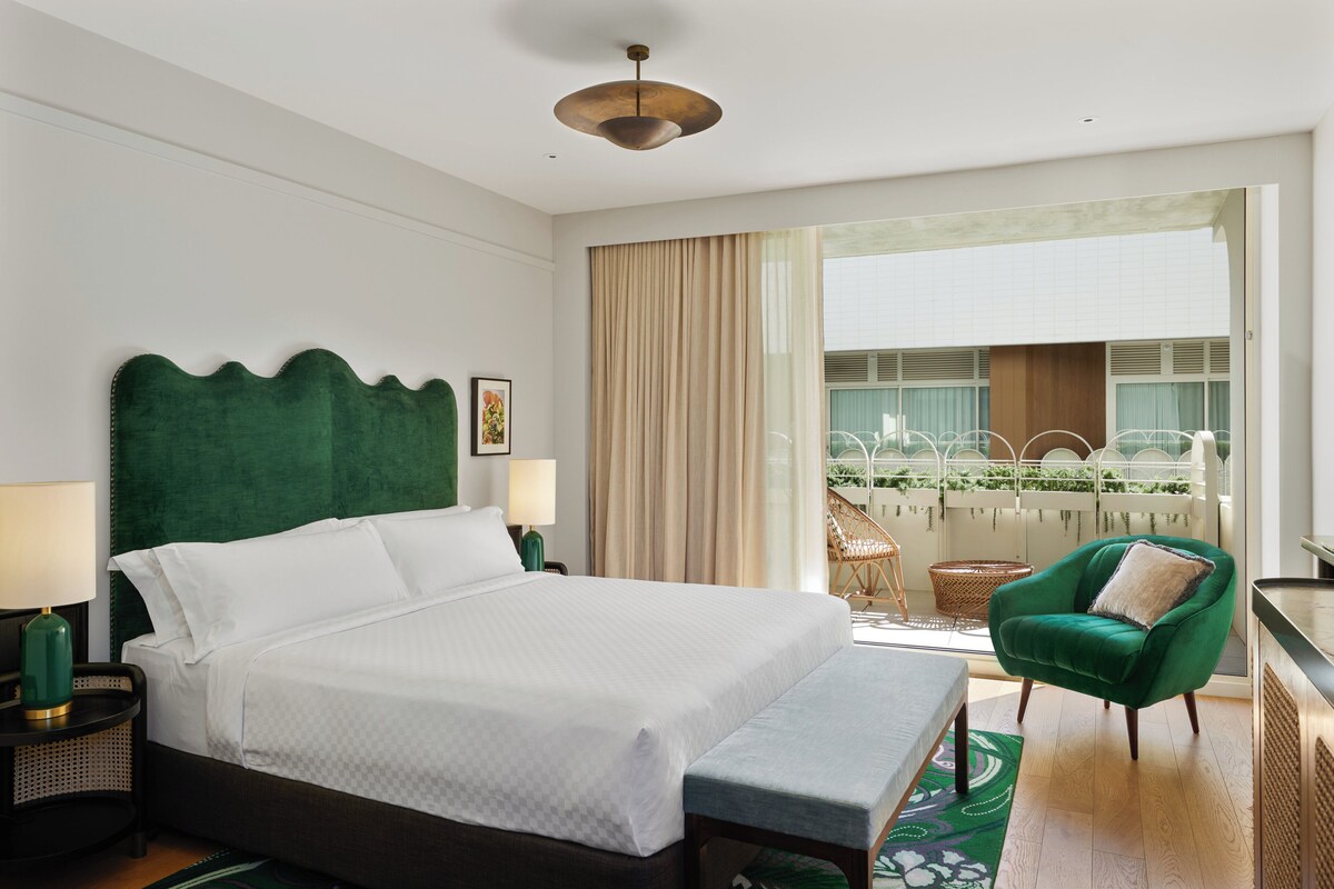 A hotel room with a large bed, a green armchair, a balcony with a view of greenery, and a ceiling light.