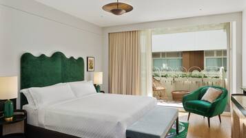 Habitación de hotel con una cama grande, un sillón verde, un balcón con vistas a zonas verdes y una lámpara en el techo.