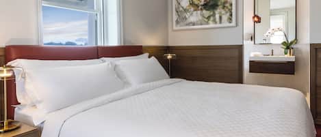 A hotel room with a bed, a nightstand, a lamp, a window with a view, a wall-mounted mirror, and a framed picture.