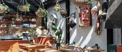 An outdoor seating area with wooden tables and chairs, hanging baskets with plants, and a bar area with bottles and glasses.