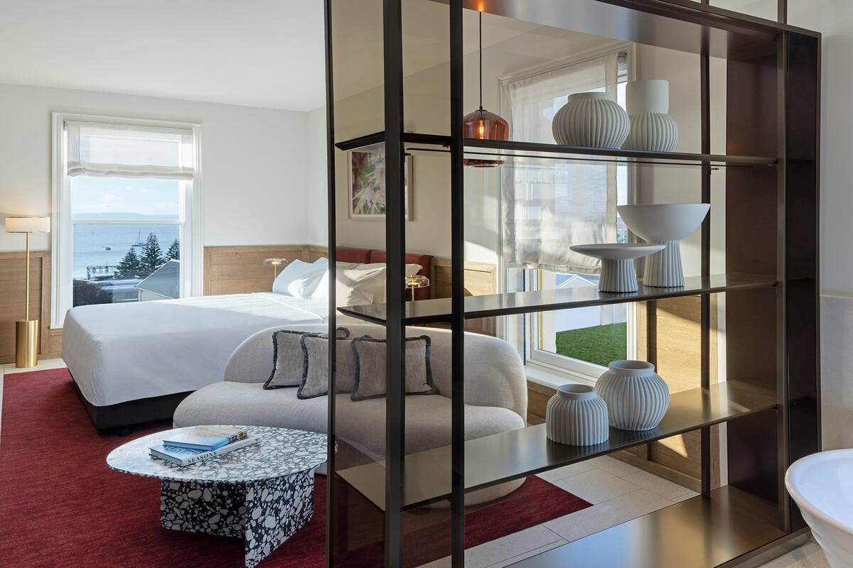Een moderne hotelkamer met een groot bed, een zithoek met een salontafel, een glazen scheiding met decoratieve elementen, en uitzicht op zee.